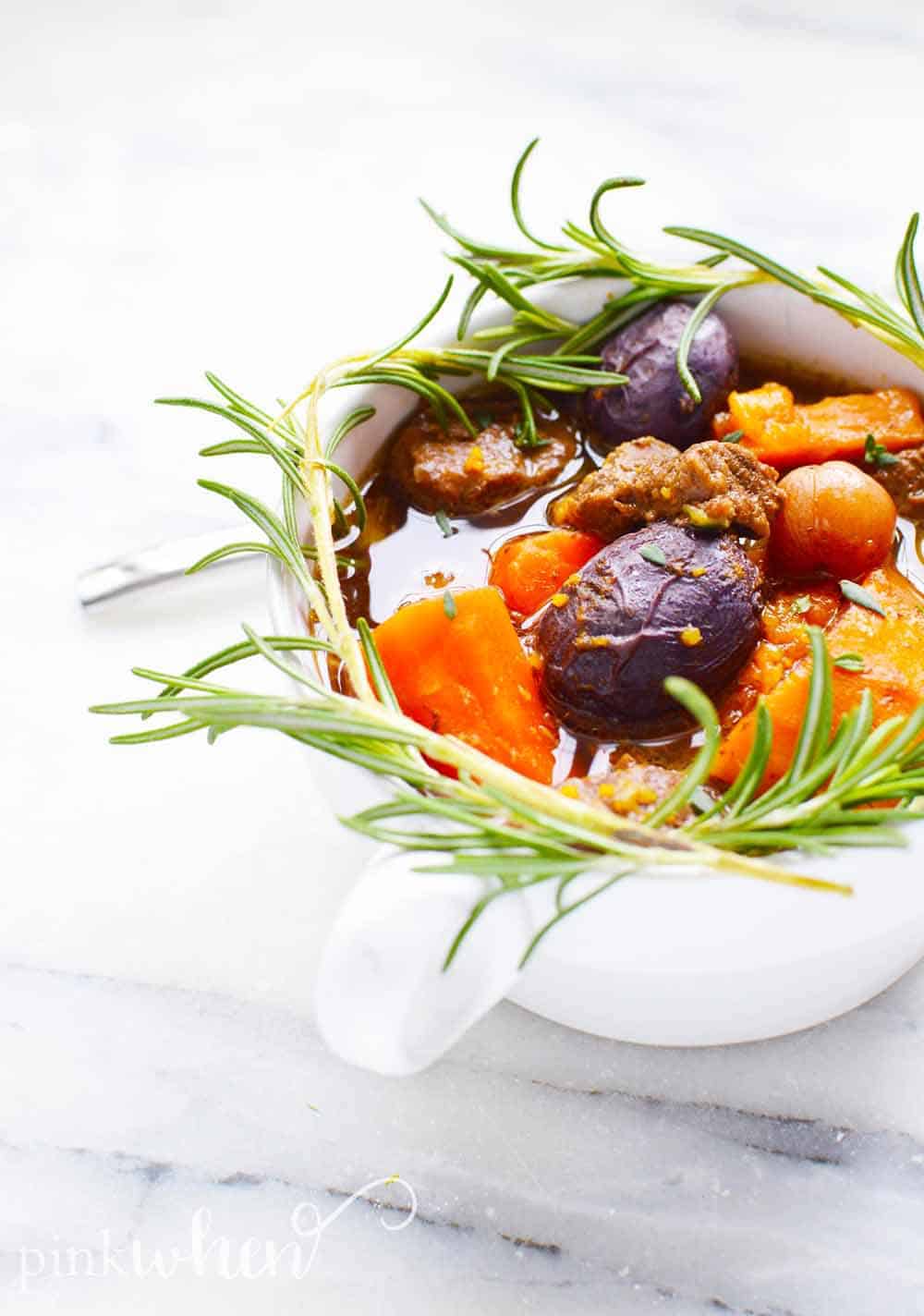 A bowl of lamb stew in a white counter with rosemary sprigs around it. 