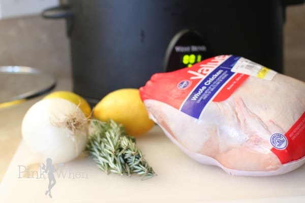 Whole chicken, onion, lemon, and rosemary sprigs on white cutting board in front of a slow cooker. 