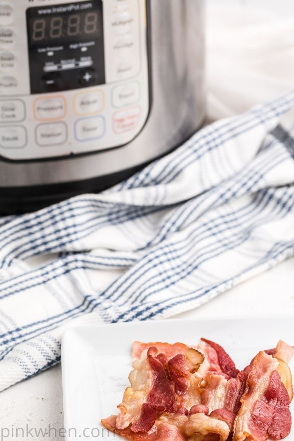 Pieces of bacon on a white plate in the foreground with an Instant Pot in the background.