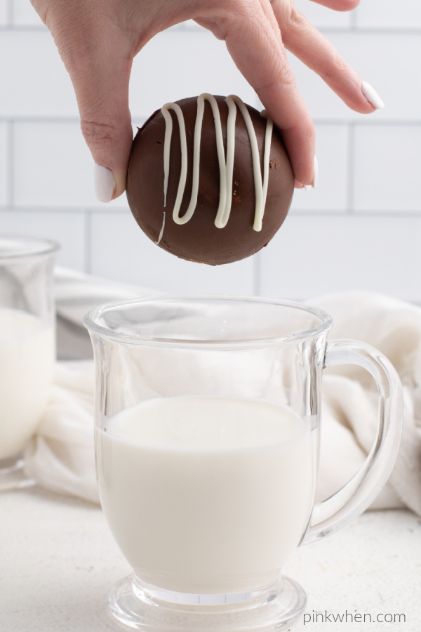 Hand holding a hot chocolate bomb about to be dropped in a glass of milk. 