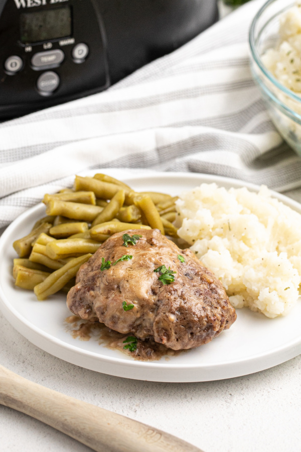 Slow Cooker Salisbury Steak on a plate with green beans and mashed potatoes with a Crock Pot in the background. 