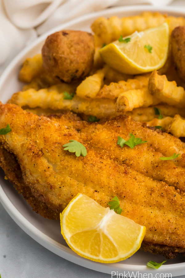 Air fried Catfish fillets on a white plate with lemon wedges, french fries, and hush puppies. 