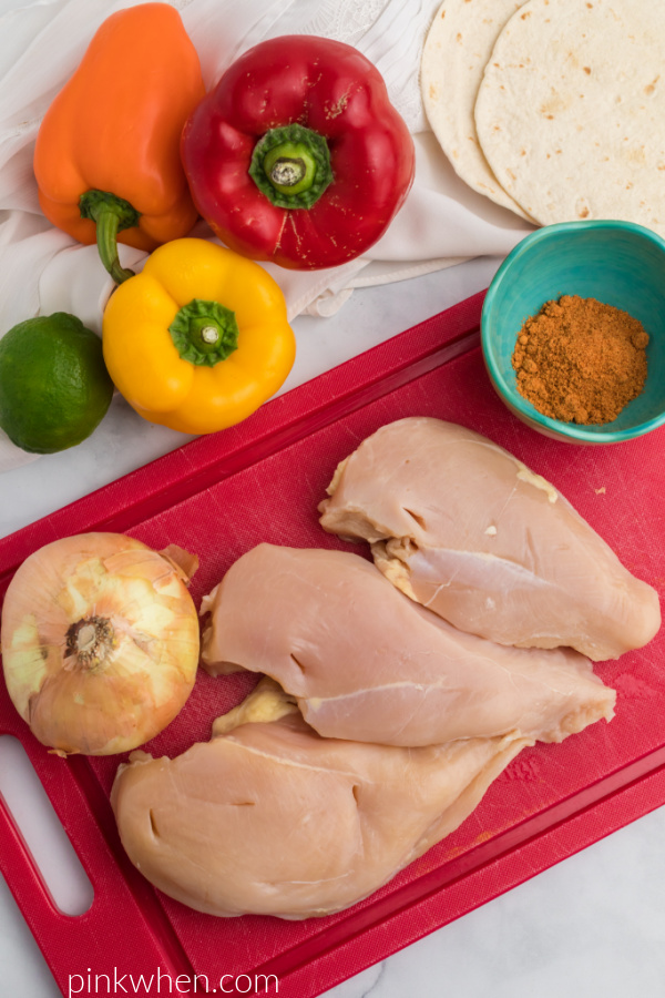 chicken breasts on a cutting board and taco seasoning, peppers, lime, and tortillas surrounding the chicken on the cutting board. 