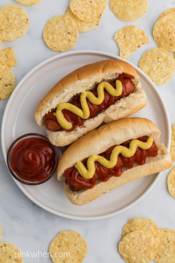 Hot Dogs in buns that were made in the air fryer, on a white plate, topped with mustard and ketchup, and surrounded by chips. 