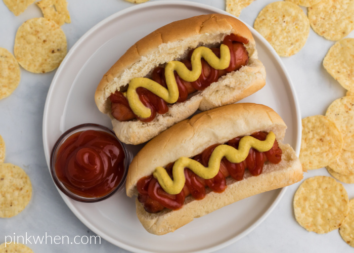 Hot dogs covered in mustard and ketchup that were made in the air fryer - served on a white plate with extra ketchup and surrounded by chips. 