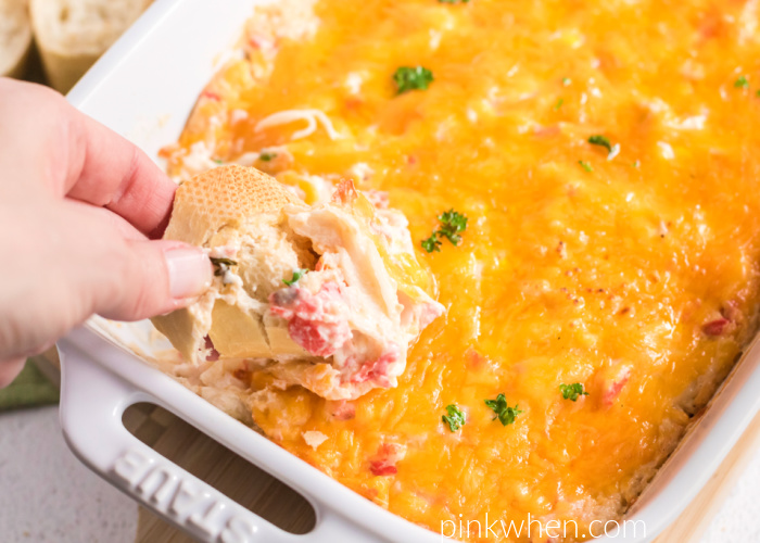 Hot crab dip in a casserole dish with a  hand scooping some cheesy crab dip with bread. 