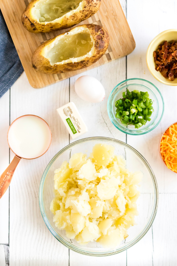 Ingredients needed for twice baked potatoes to be made - pototo pulp in a bowl, potato shells with crispy skin on a cutting board, and ingredients to add to the potato flesh on the table ready to be added. 