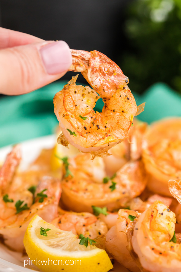 Piece of air fryer lemon pepper shrimp ready to be eaten - held by a hand over a plate of shrimp. 
