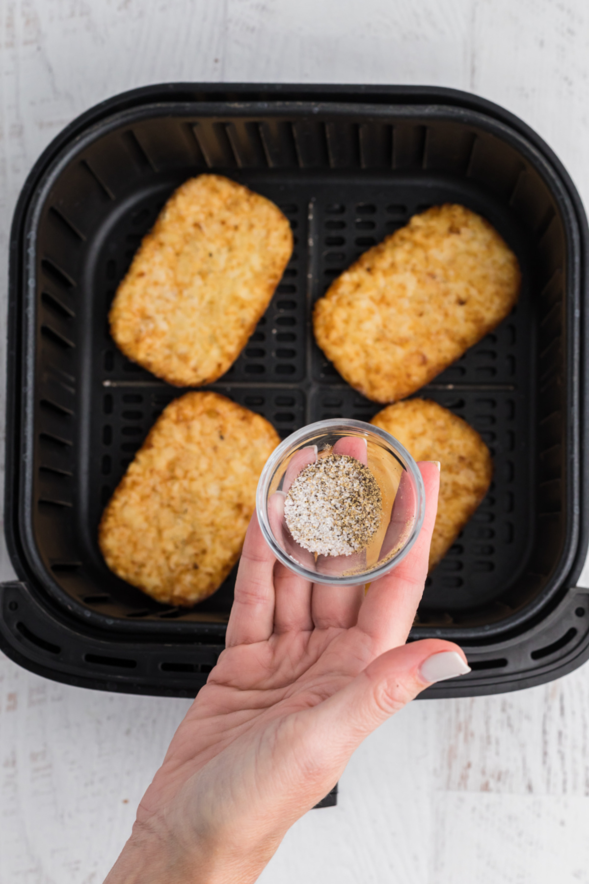 frozen hash brown patties in the basket of the air fryer with a hand holding salt and pepper to season them with before cooking. 