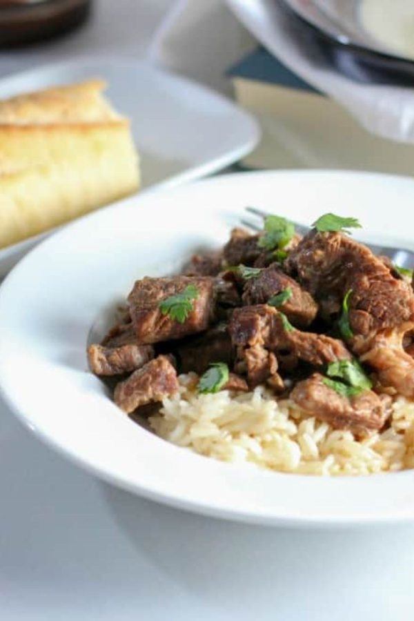 Juicy beef tips in a white bowl over rice, served with bread on a white table. 