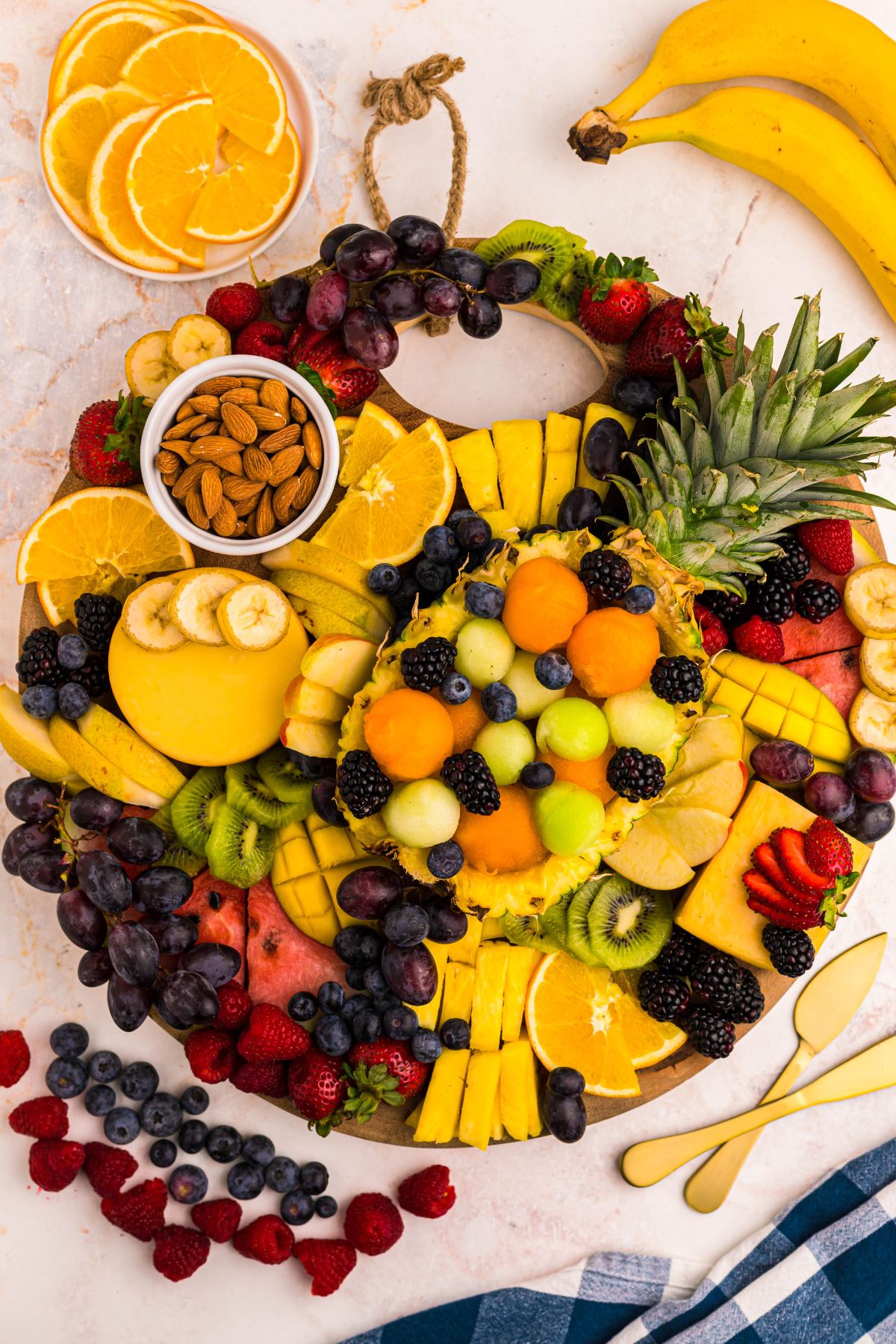 Various fruits being added to charcuterie board with extra pieces of fruit on the table to fill open spots. 