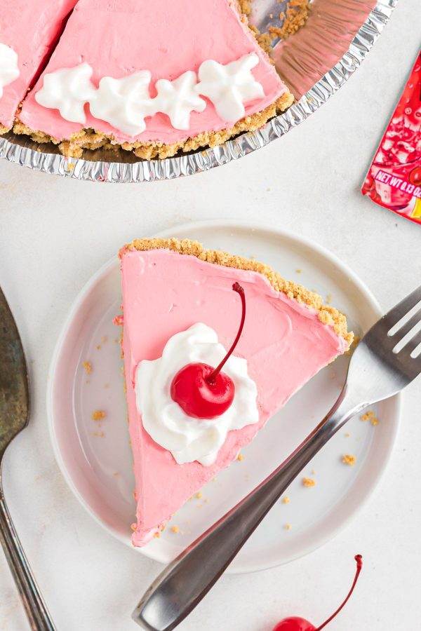 Pink cherry Kool Aid pie sliced on a small white plate topped with cool whip and cherry. 