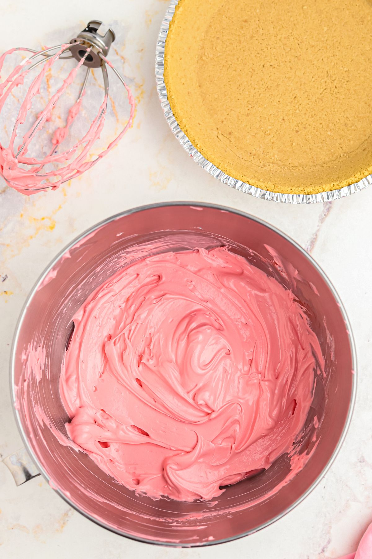 Ingredients mixed in a silver mixing bowl on a white marble table next to a graham cracker crust.