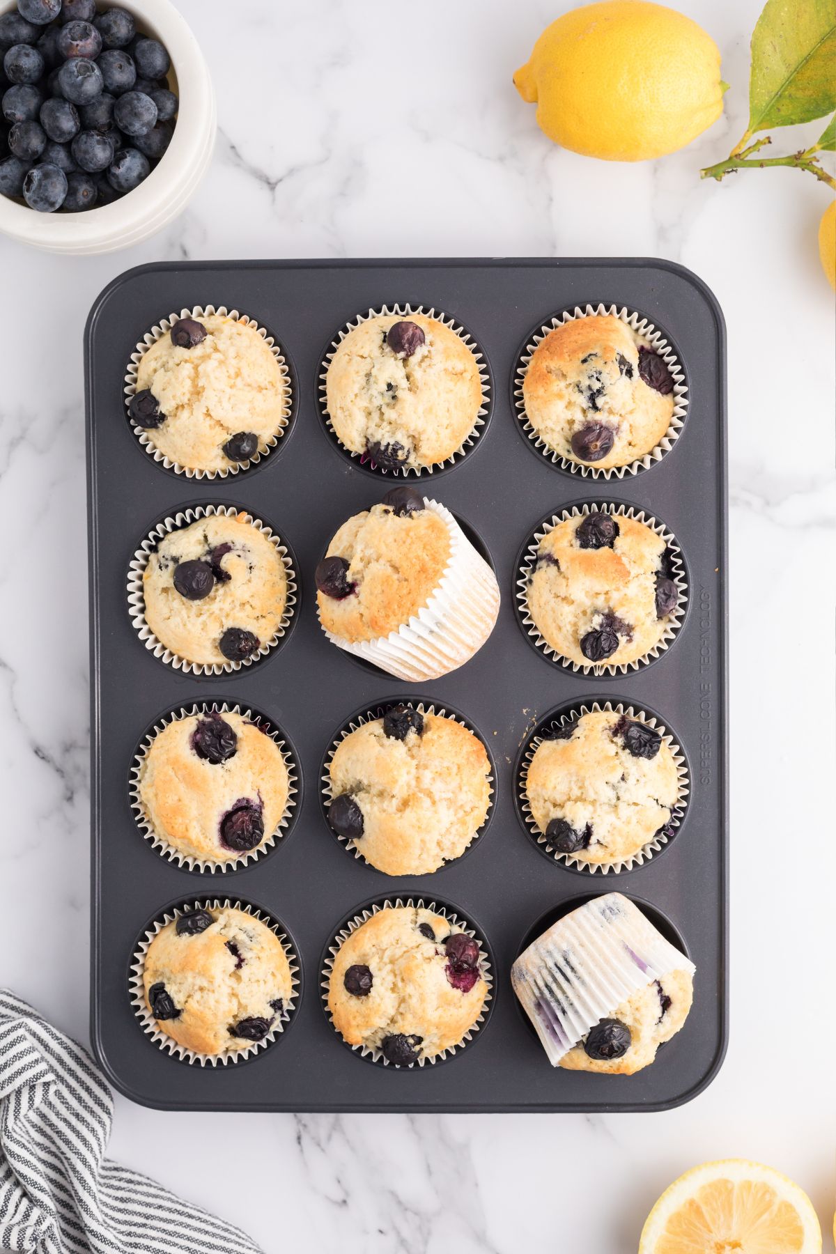 Golden baked lemon blueberry muffins in a muffin tin on a white marble table.