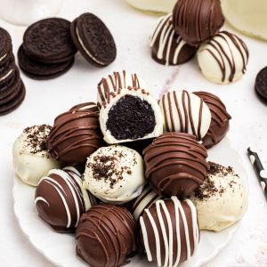 White chocolate and milk chocolate coated Oreo balls stacked on a white plate in front of milk and Oreo cookies.