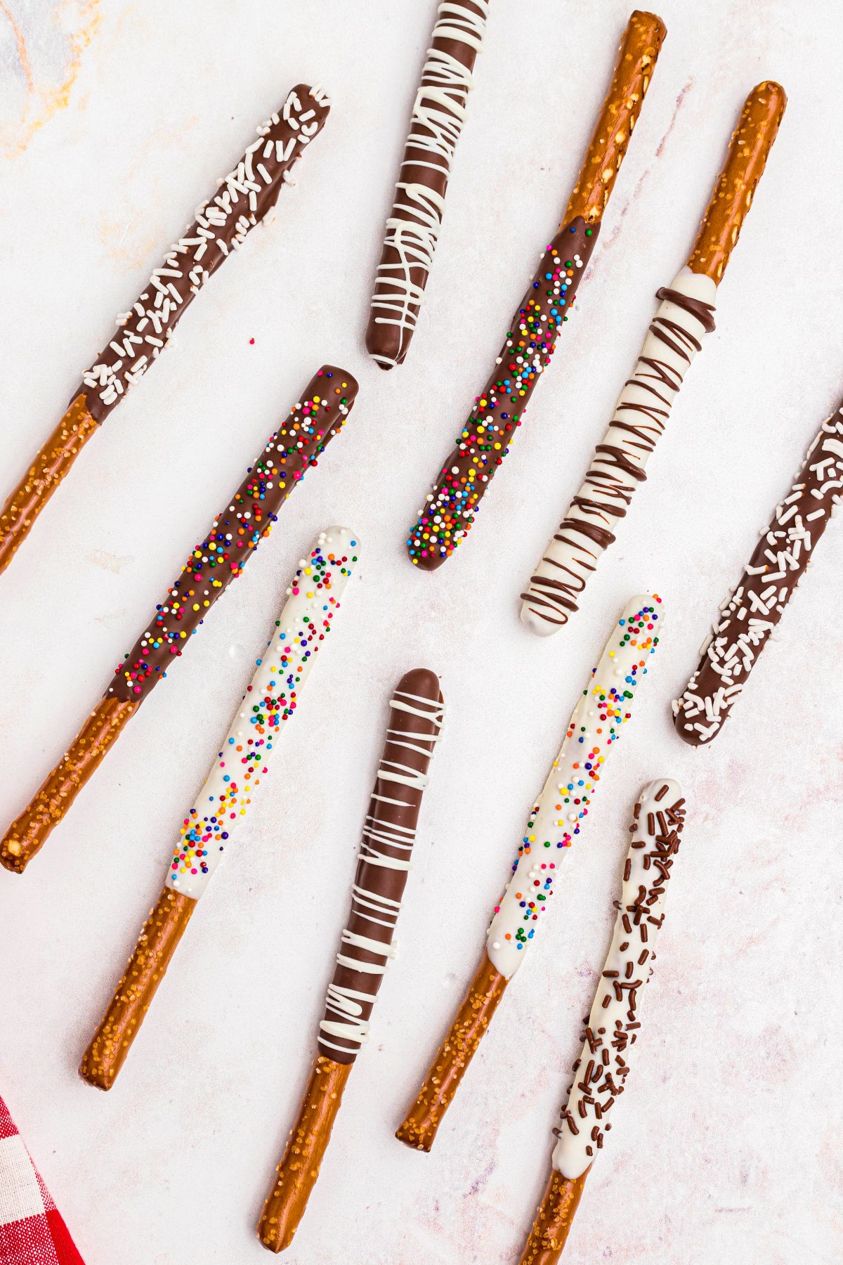 Pretzel rods drying on a marble table after being dipped in chocolate and white chocolate and coated in sprinkles. 