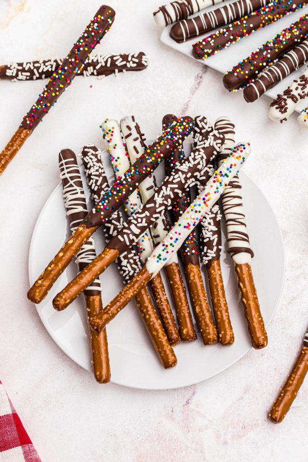 Chocolate and white chocolate pretzel rods with sprinkles and drizzles stacked on a white plate. 
