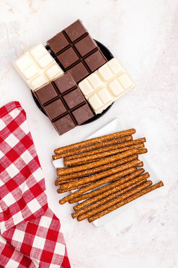 Blocks of chocolate on a black plate and pretzel rods on a white square plate. 