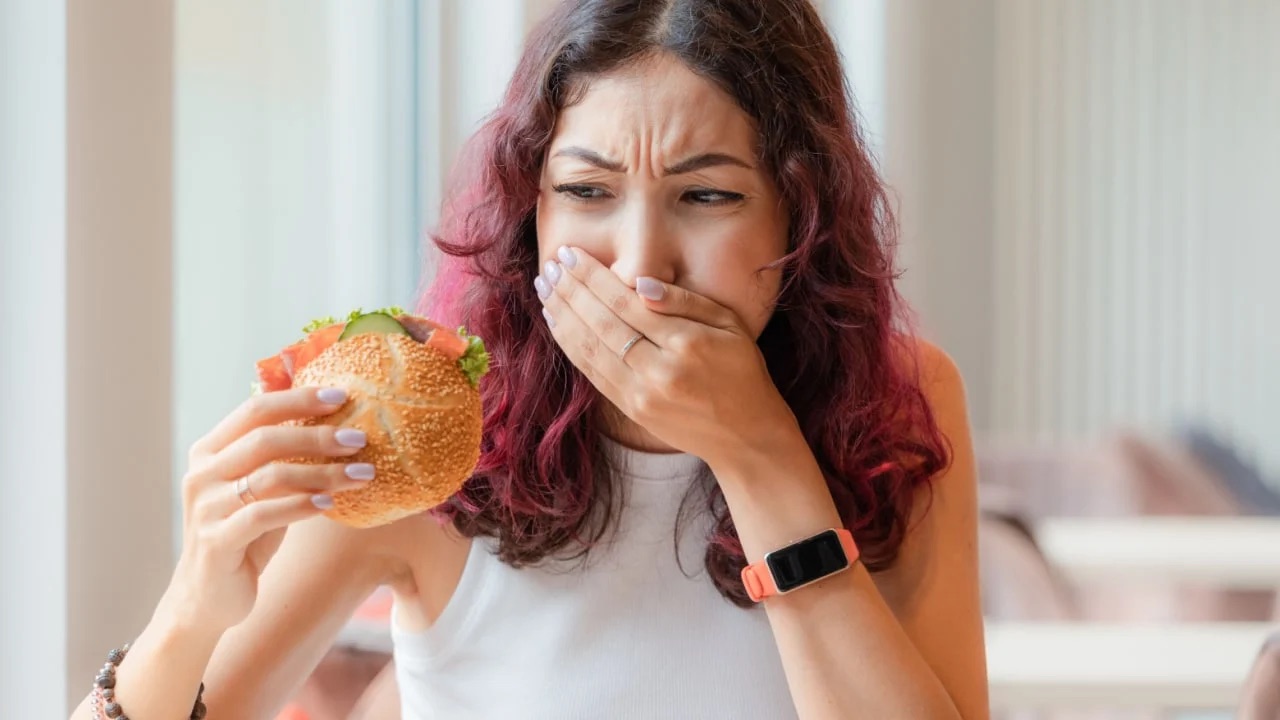 Woman holding a burger with hand over her mouth.