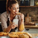Young happy woman text messaging on mobile phone and eating Gluten-Free-Bread in the kitchen.
