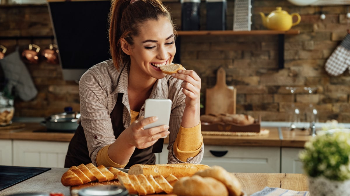 Young happy woman text messaging on mobile phone and eating Gluten-Free-Bread in the kitchen.