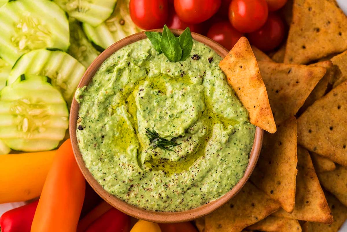 A bowl full of green goddess dip served with chips and fresh vegetables.