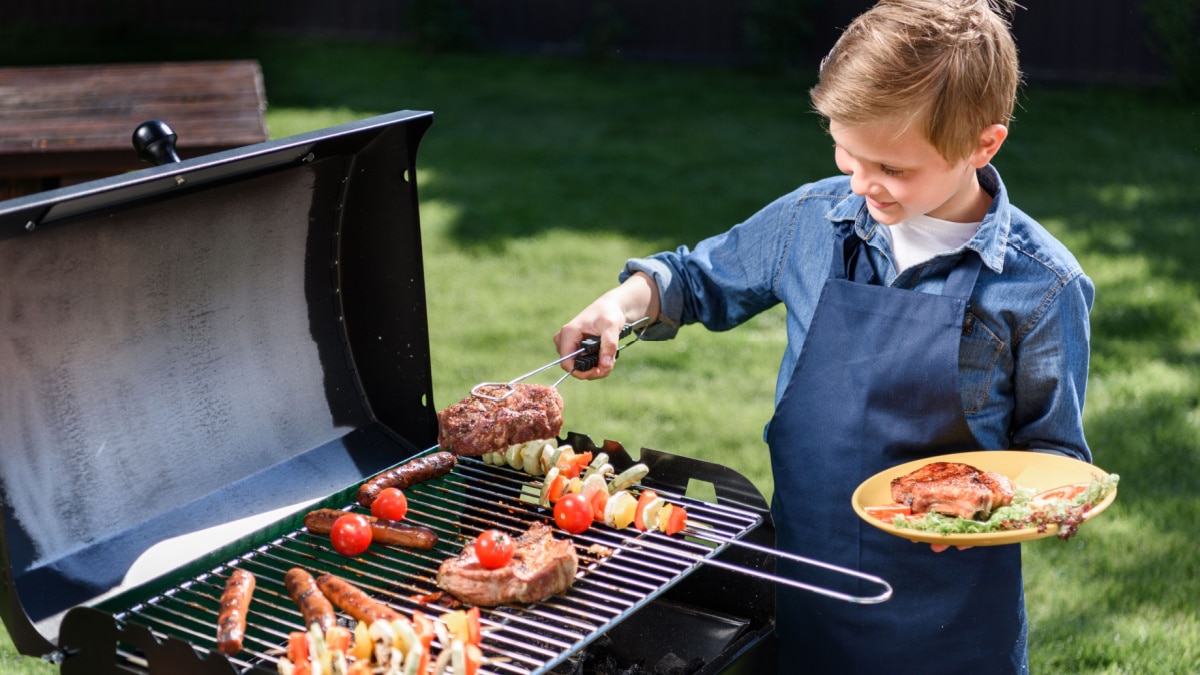 Smiling boy in apron preparing tasty steaks on a barbecue grill outdoors.