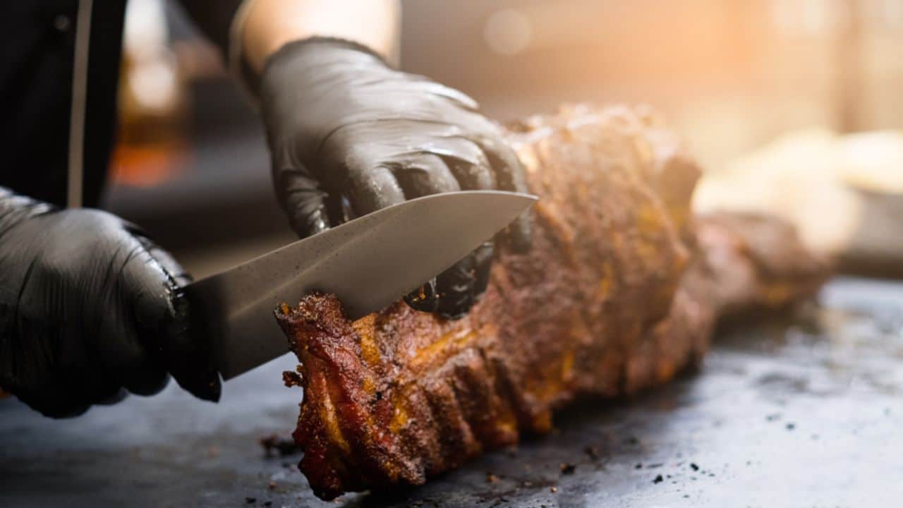 Chef in black cooking gloves using knife to cut smoked pork ribs.