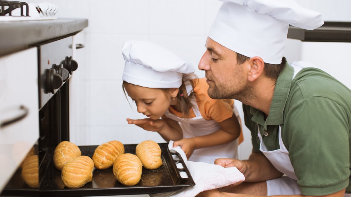 Father with daughter take some rolls on a baking tray out of oven.