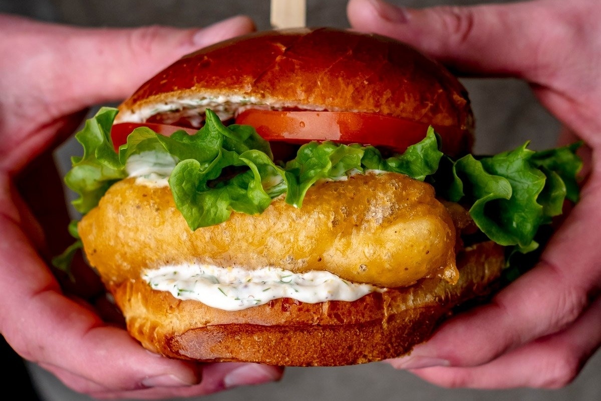 A person holding up a fish burger with lettuce and tomatoes, served on a bun.