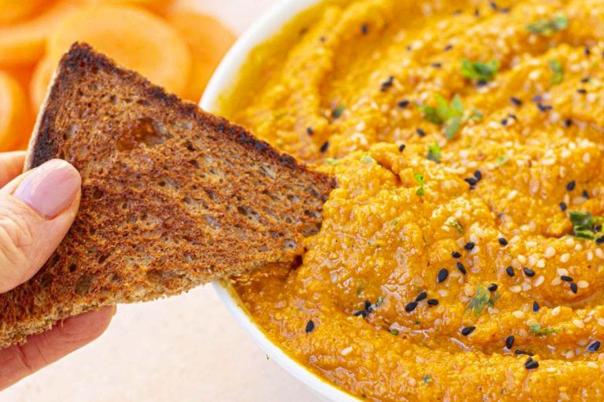 A person is enjoying a delicious game day dip by taking a piece of bread out of a bowl of carrot hummus.