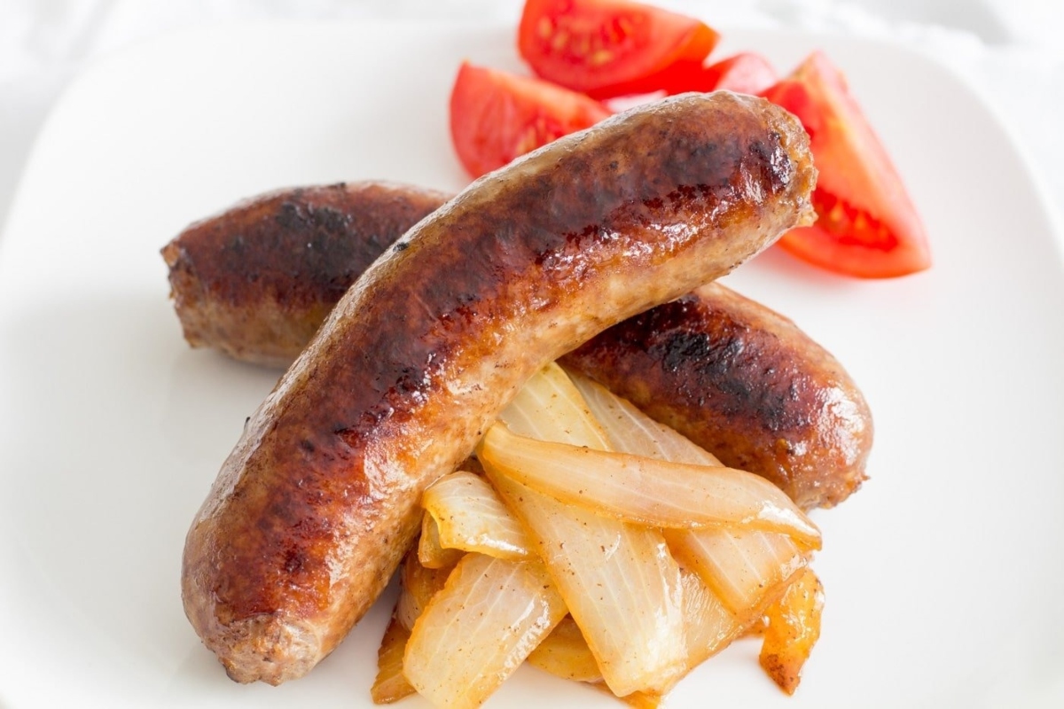 Sausages served on a bun, topped with onions and tomatoes, presented on a white plate.