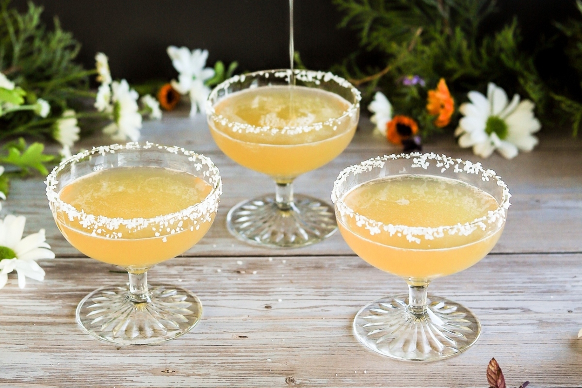 Three cocktails in margarita glasses on a wooden table with flowers.