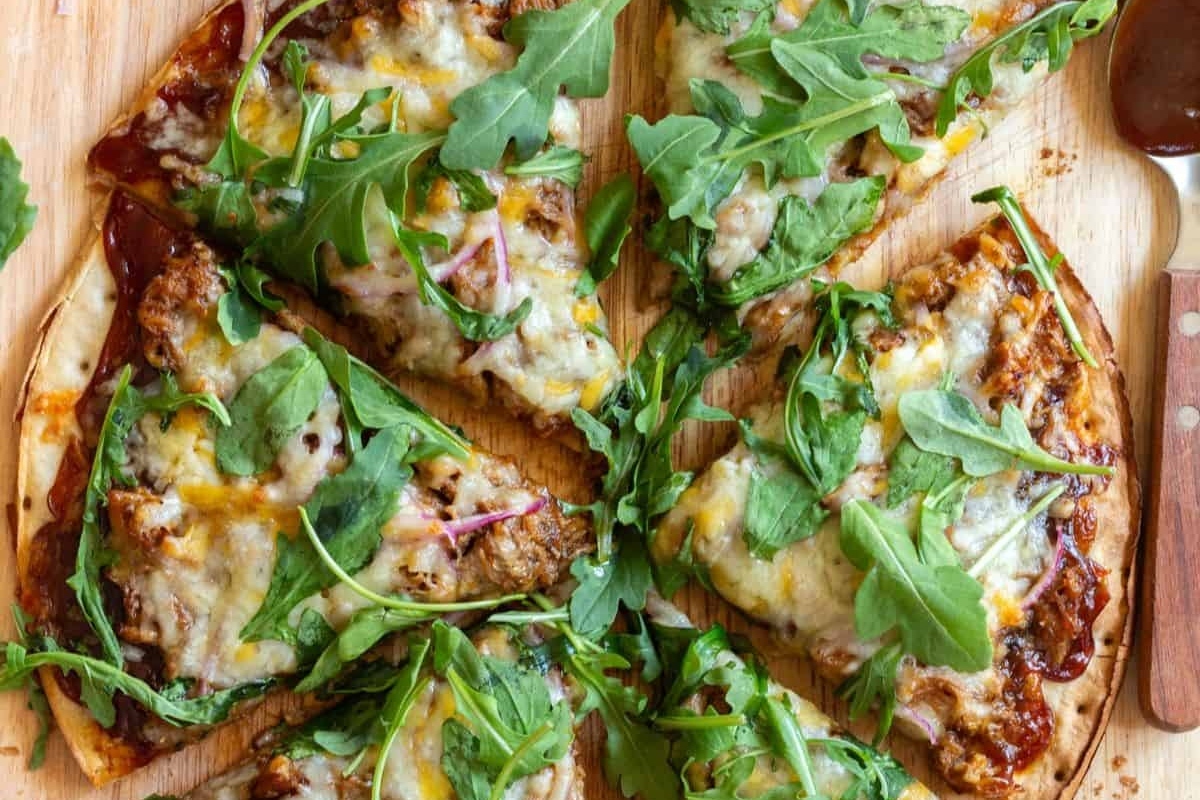 A pork pizza with fresh greens on a wooden cutting board.