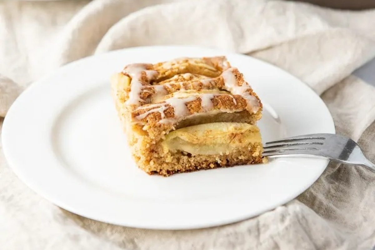 A slice of apple cinnamon roll cake on a plate with a fork. The cake is made with fresh apple and infused with cider flavor.