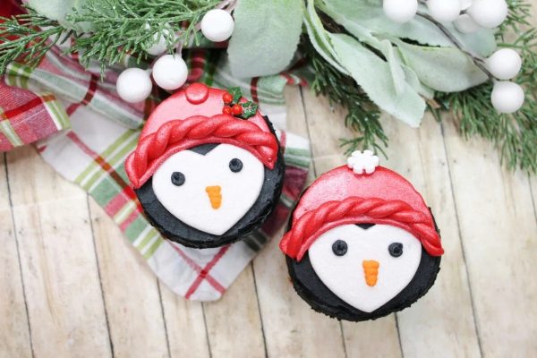 Two Christmas penguin cupcakes with red hats on a wooden table.