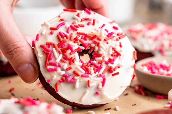 A person holding a delicious red velvet donut with sprinkles on it.