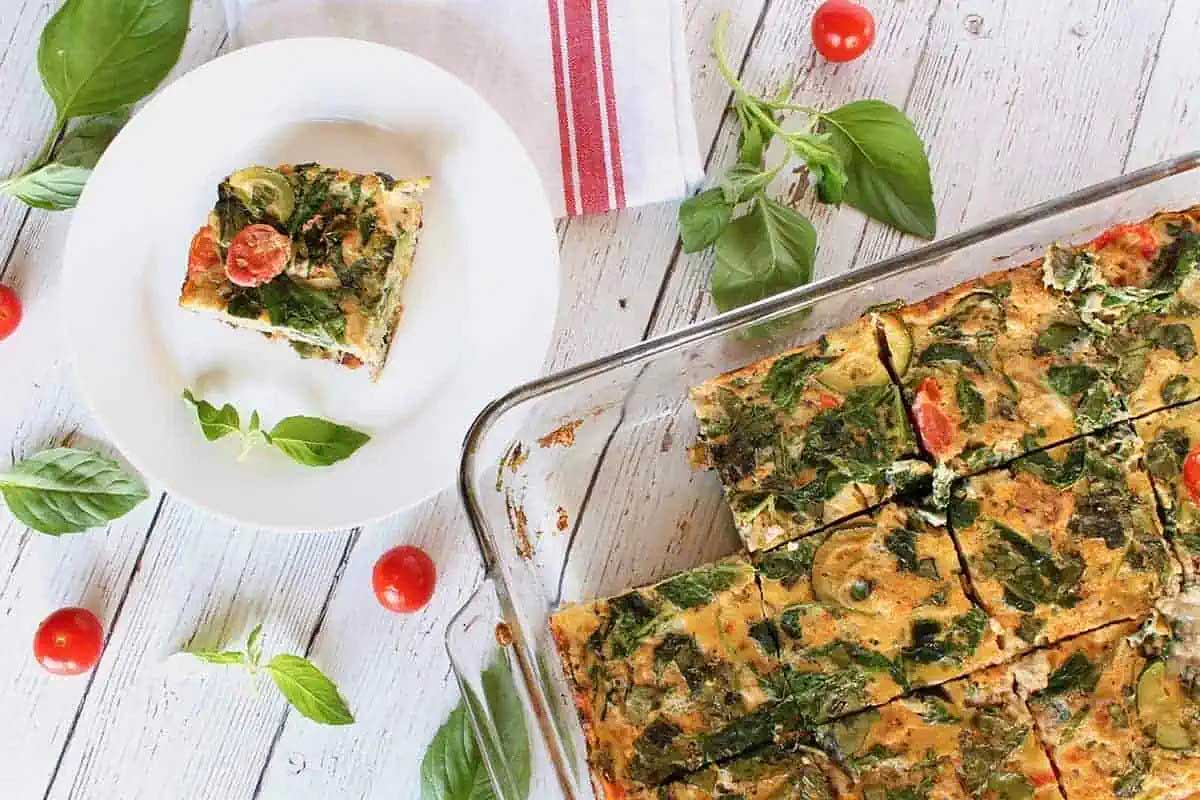 A slice of quiche with spinach and tomatoes on a white plate.