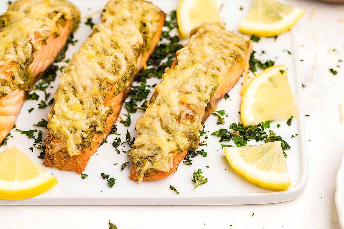 Three salmon fillets on a white plate with lemon wedges, drizzled with a flavorful pesto sauce.