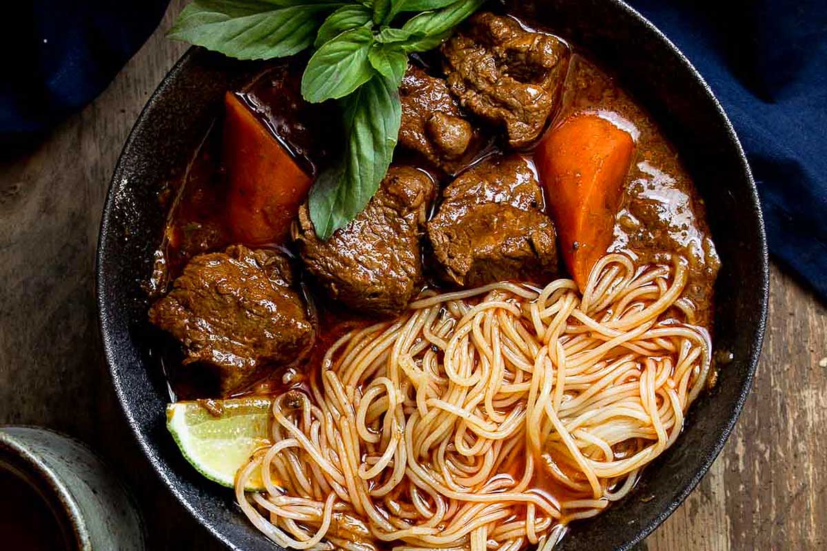 A bowl of beef stew with noodles and carrots on a wooden table.