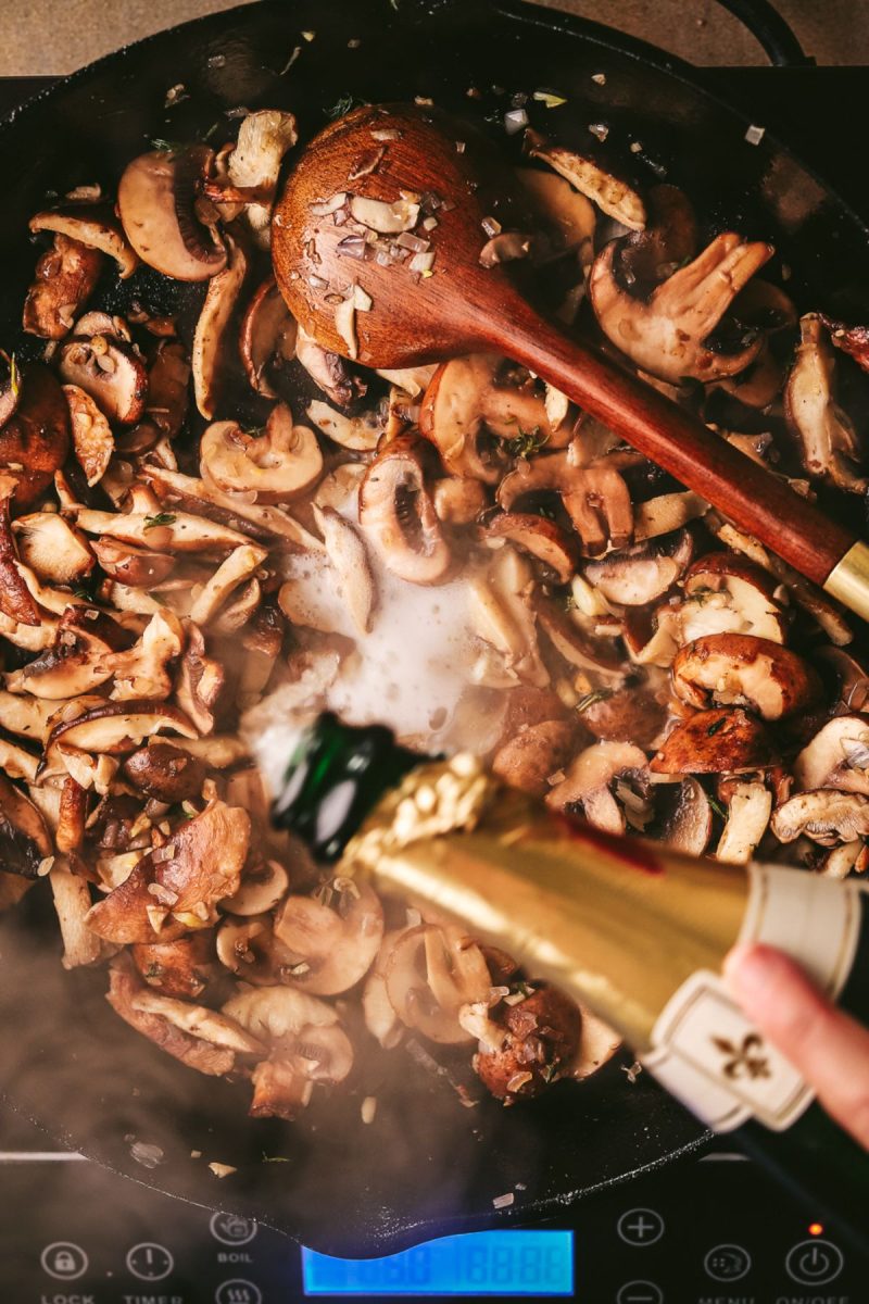 A person pouring champagne into a pan with mushrooms.