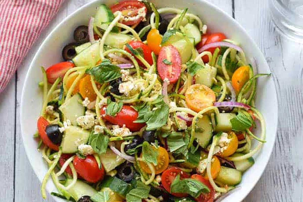 Zucchini salad in a white bowl with tomatoes and olives.