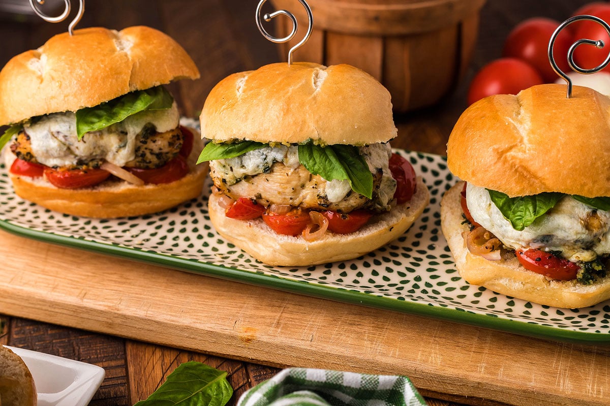 Three chicken sliders served on a bun with tomatoes and basil on a plate.
