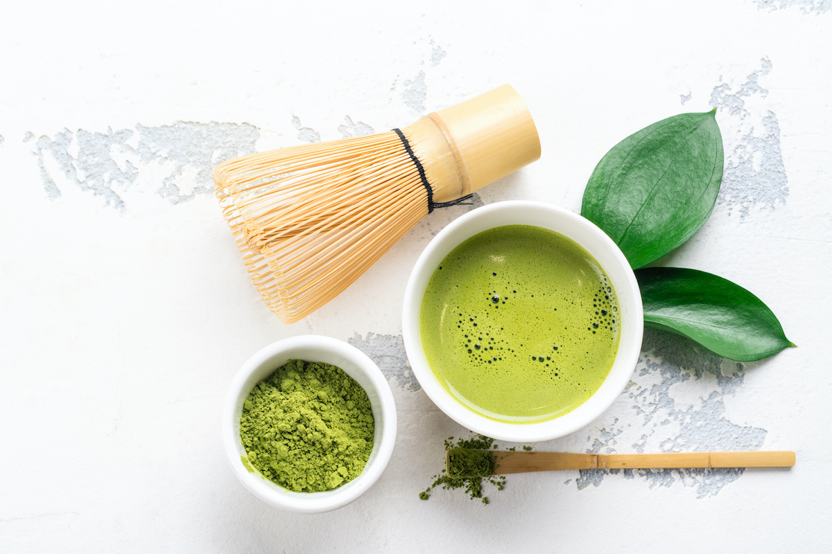 A bowl of matcha powder and a bamboo whisk on a white background.