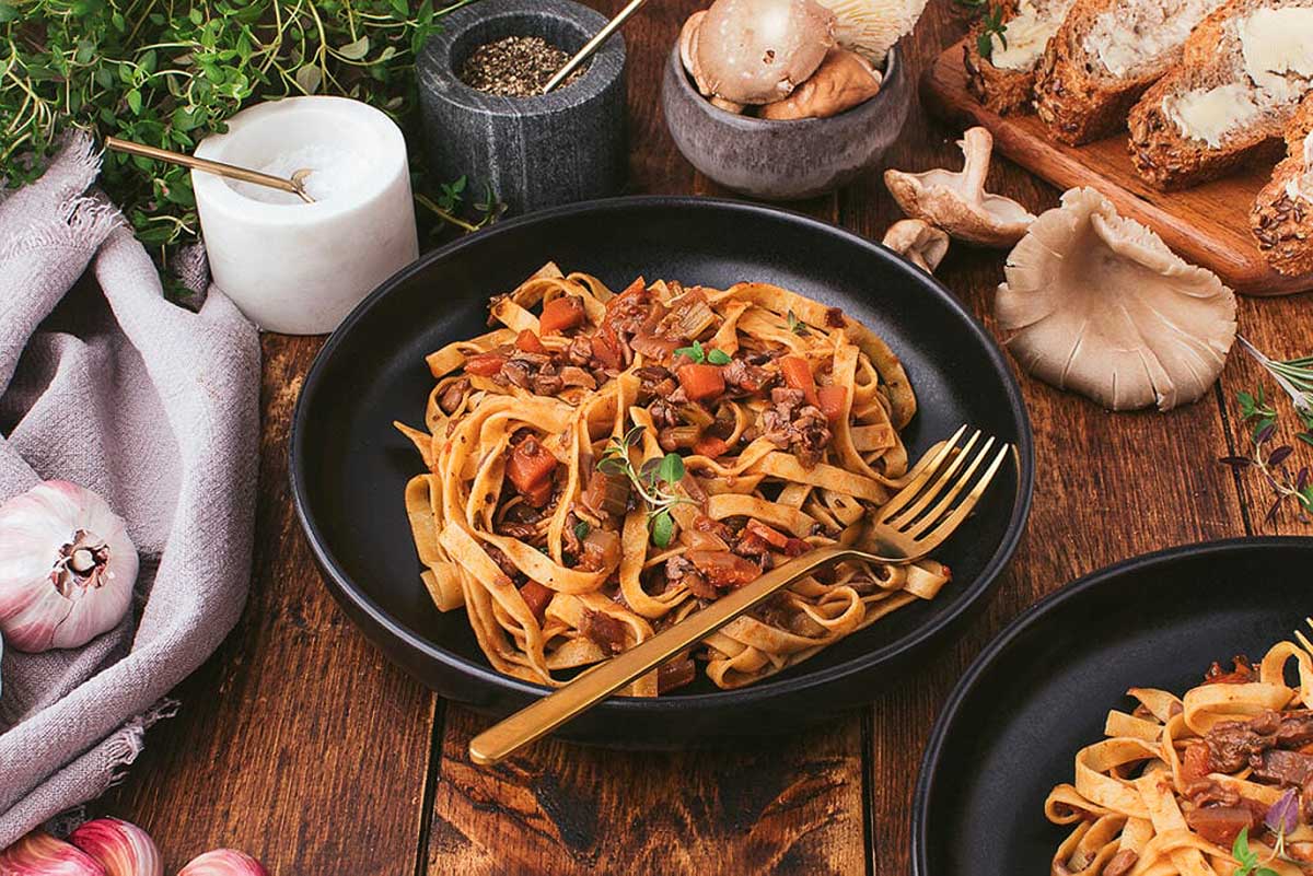 Two amazing bowls of pasta with meat, vegetables, and mushrooms on a wooden table.