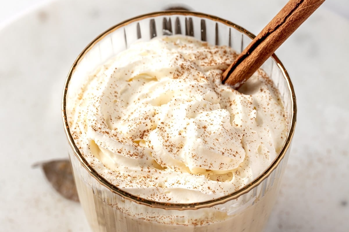 A glass of eggnog topped with whipped cream and garnished with cinnamon sticks.