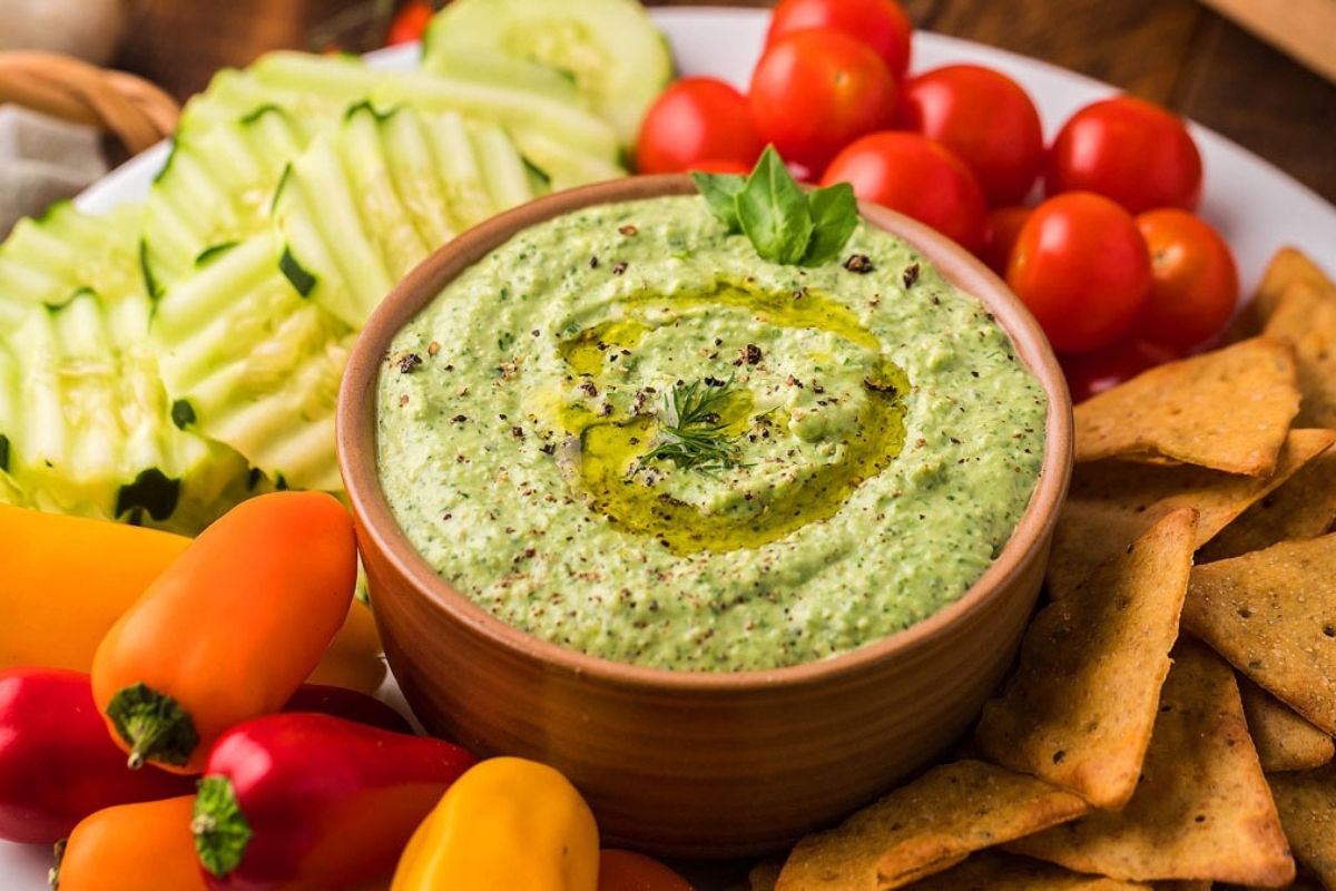 A bowl of green spinach pesto hummus with crackers and vegetables.