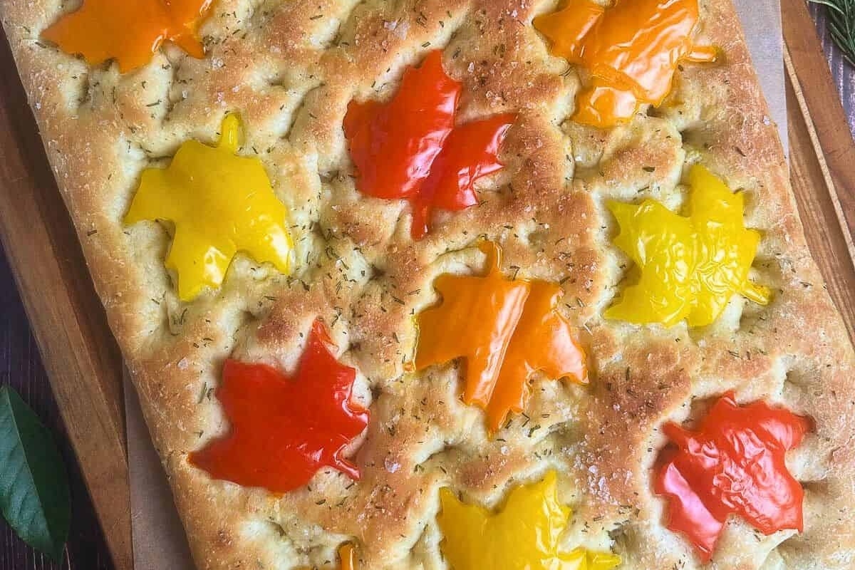 A mouthwatering focaccia topped with peppers and fresh leaves on a rustic cutting board.