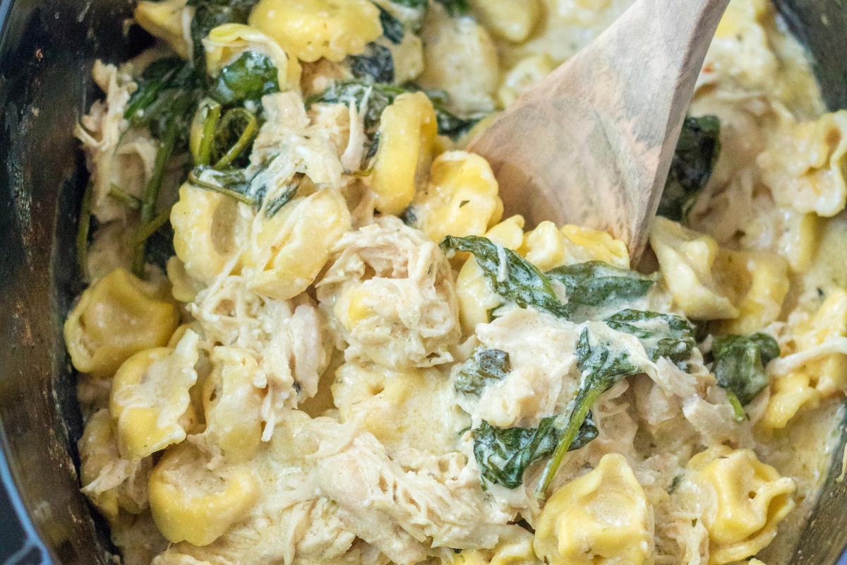 Chicken and spinach tortellini recipe in a slow cooker with a wooden spoon.