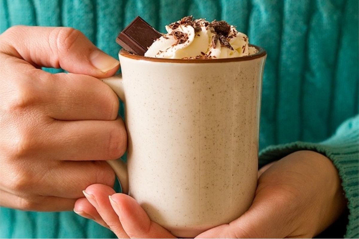 A person holding a mug of hot chocolate with whipped cream and chocolate.